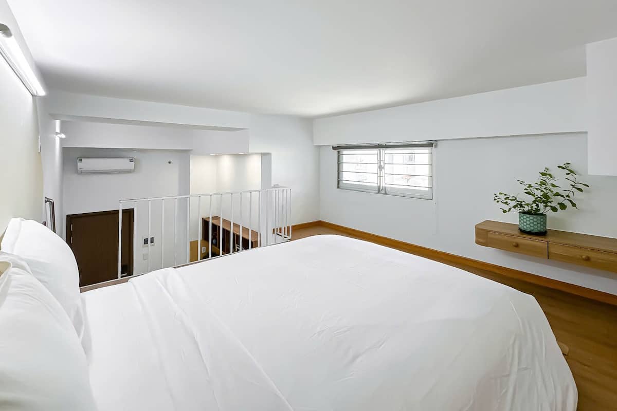 A spacious, well-lit room with a large bed, a window, a staircase, and a small plant on a shelf.