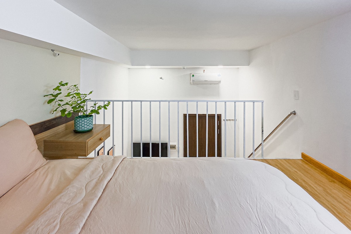 A bedroom with a bed, a nightstand with a plant, a wooden headboard, a white wall, a white ceiling, a white railing, and a wooden staircase.