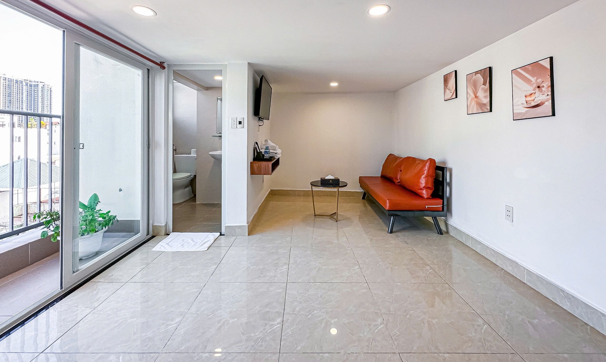 A modern living room with a glass door leading to a balcony, a red leather sofa, a small table, and a TV mounted on the wall.