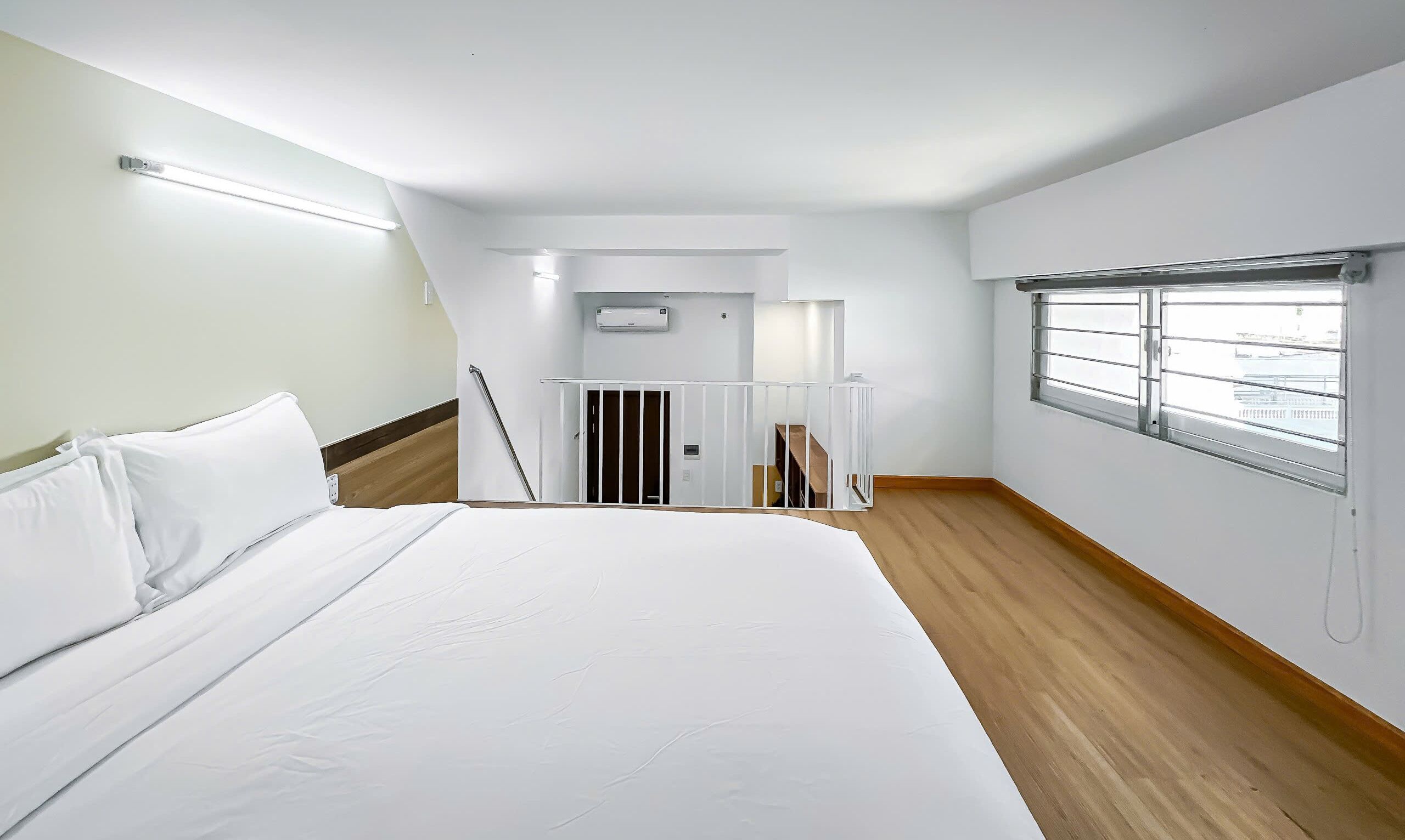 Habitación de hotel moderna con una cama grande, suelo de madera, ventana con persianas, pared blanca y una lámpara en el techo.