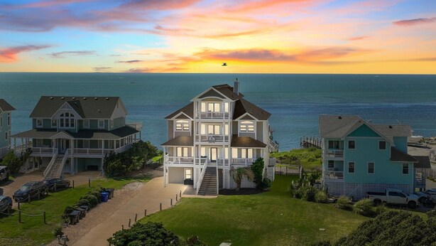 Une grande maison à plusieurs étages, dotée d’un porche à l’avant et d’un escalier menant à l’entrée, située au bord d’un plan d’eau, avec un coucher de soleil en arrière-plan.