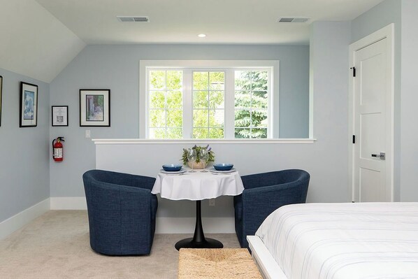 A room with a bed, two blue chairs, a small table with a white tablecloth, a window with a view of trees, and a fire extinguisher on the wall.