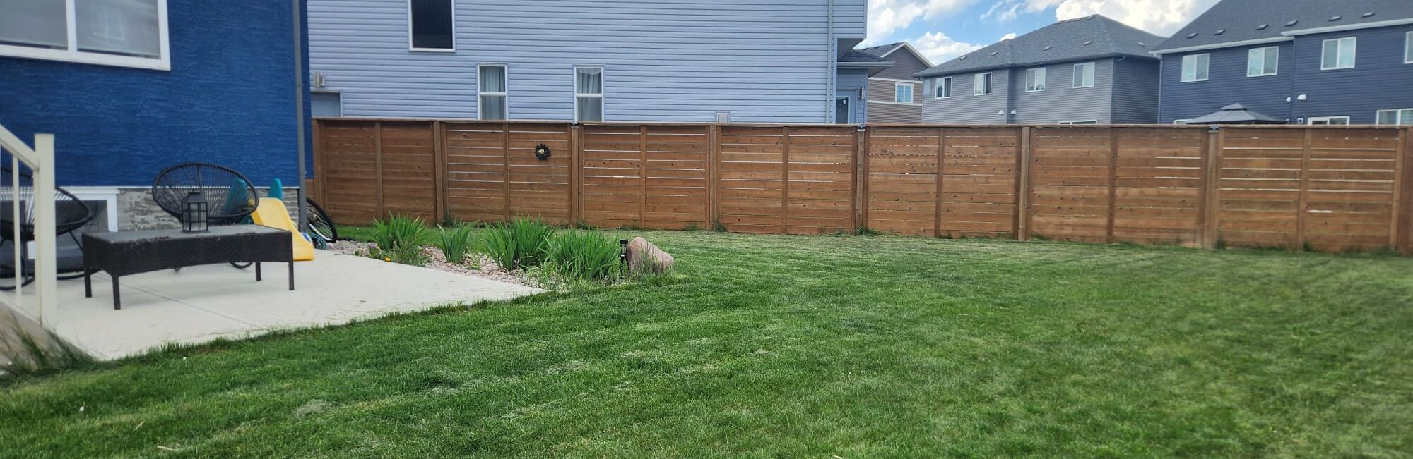 A backyard with a wooden fence, a patio with outdoor furniture, and a lawn.