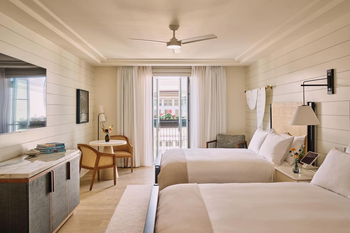 A hotel room with two beds, a desk, a chair, a small table, a lamp, and a view of the outside.