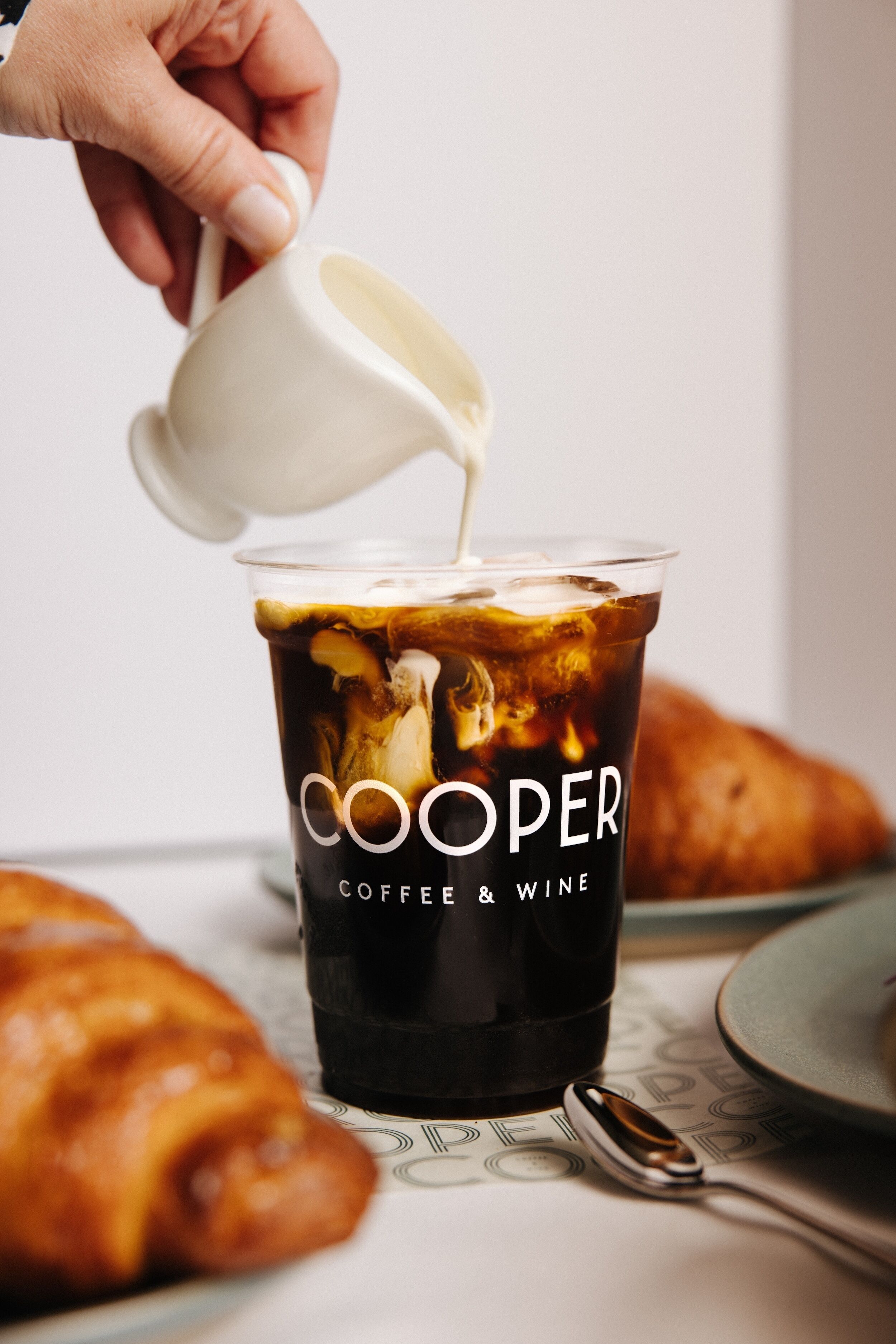 Una mano vertiendo leche en un vaso de café helado, con el texto 'COOPER COFFEE & WINE'.