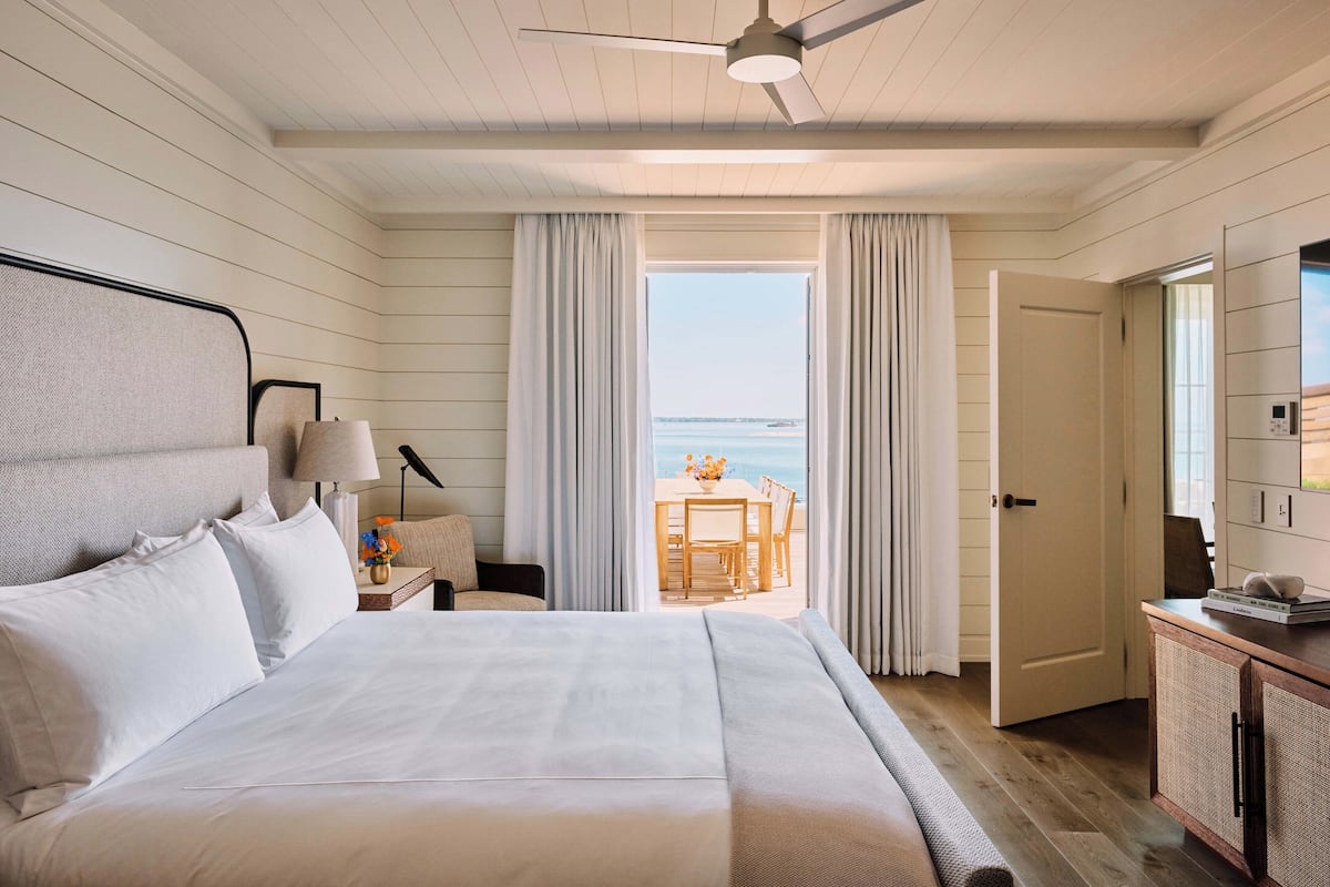 A bedroom with a bed, a nightstand, a lamp, a view of the ocean, and a door leading to a balcony.