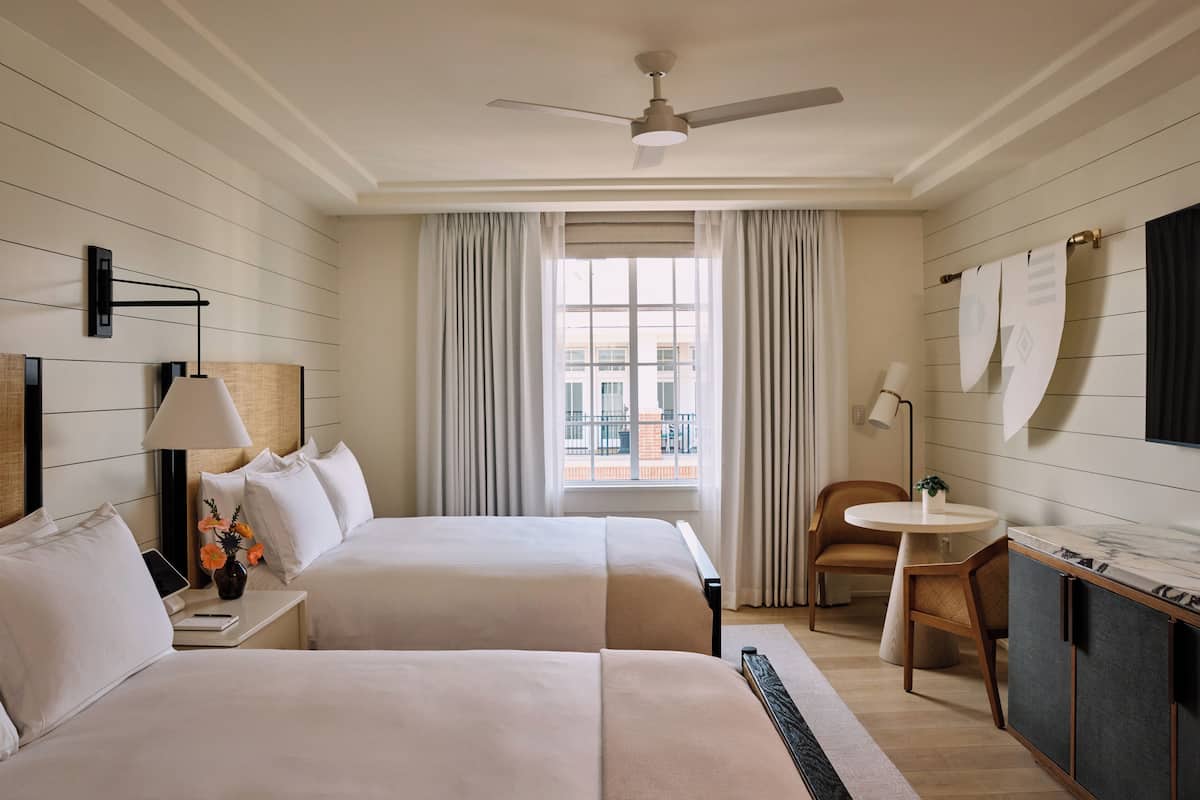 A hotel room with two beds, a desk, a chair, a small table, a ceiling fan, and a window with curtains.