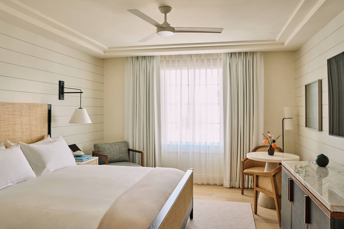 A hotel room with a bed, a desk, a chair, a ceiling fan, and a large window with curtains.