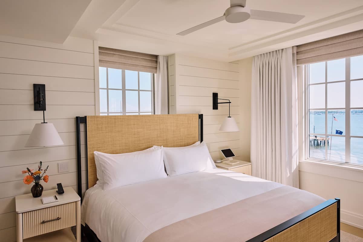 A hotel room with a bed, bedside tables, lamps, a ceiling fan, and large windows with a view of the sea.