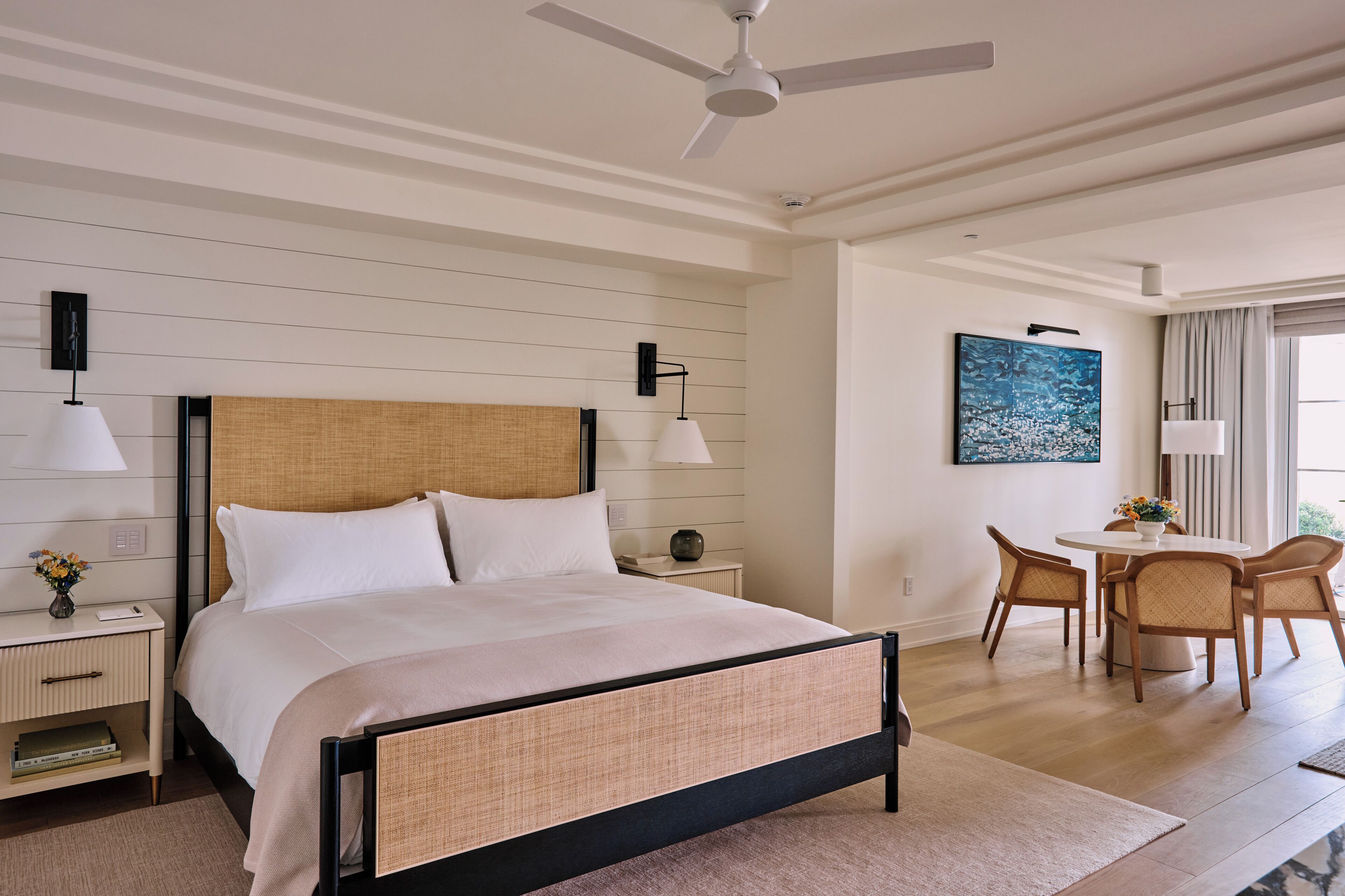 A hotel room with a bed, bedside tables, a desk, a chair, a ceiling fan, and a painting on the wall.