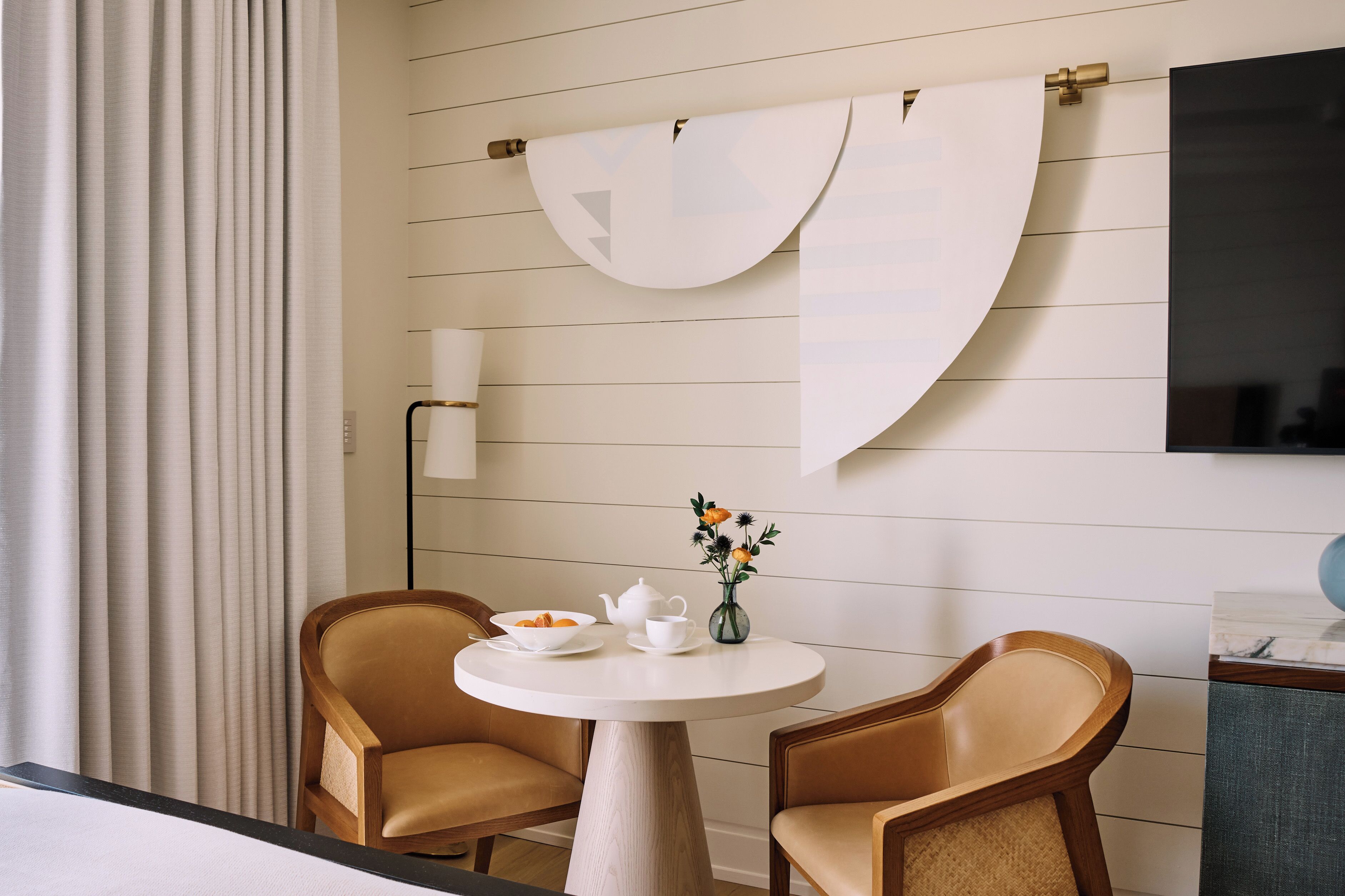 Een kamer met een ronde tafel, twee stoelen, een vaas met bloemen, een lamp en een aan de muur bevestigde televisie.