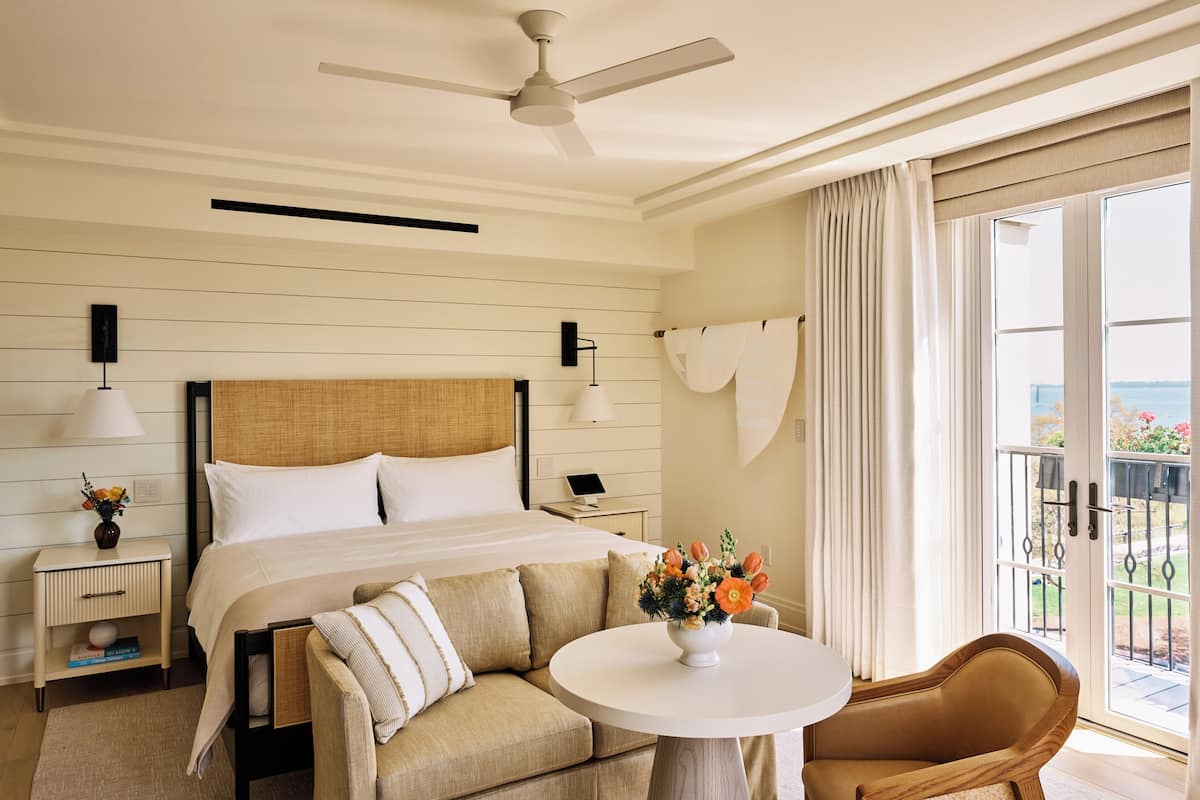 A hotel room with a bed, a round table with a vase of flowers, a sofa, a chair, a ceiling fan, and a view of the outdoors through a sliding door.
