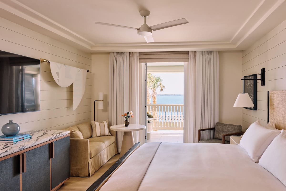 A hotel room with a bed, a sofa, a small table with a vase, a chair, a ceiling fan, and a view of the ocean.