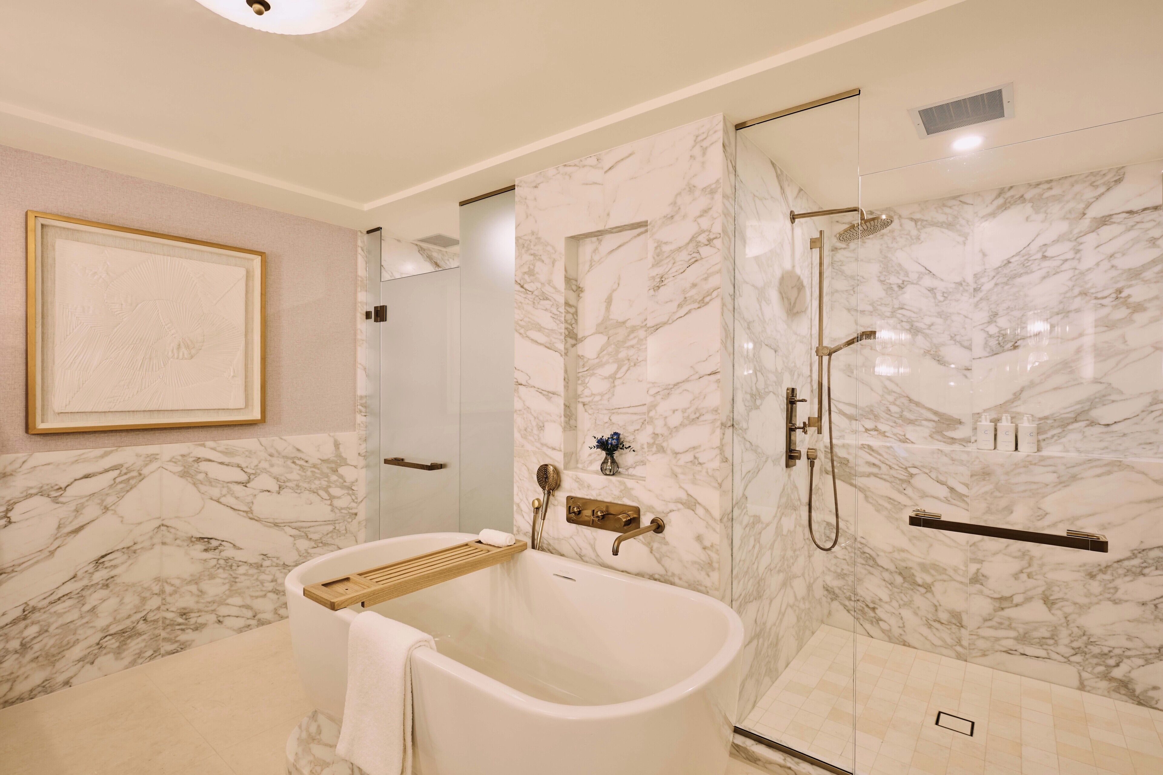 Ein Badezimmer mit freistehender Badewanne, einer gläsernen Duschkabine, Marmorwänden und einem gerahmten Kunstwerk an der Wand.
