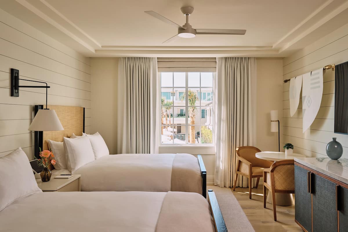 A hotel room with two beds, a desk, a chair, a window with curtains, a ceiling fan, and a view of buildings outside.