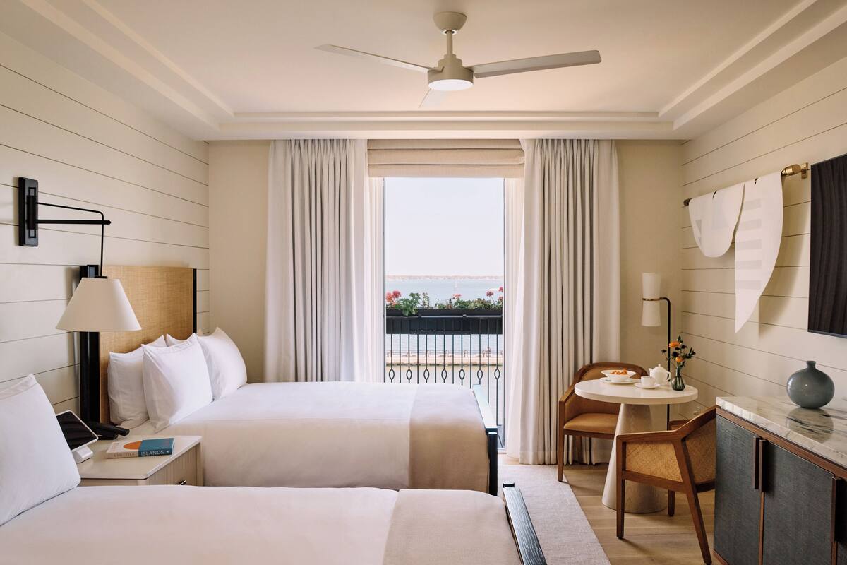 A hotel room with two beds, a desk, a chair, a small table, a lamp, a ceiling fan, and a balcony with a view.