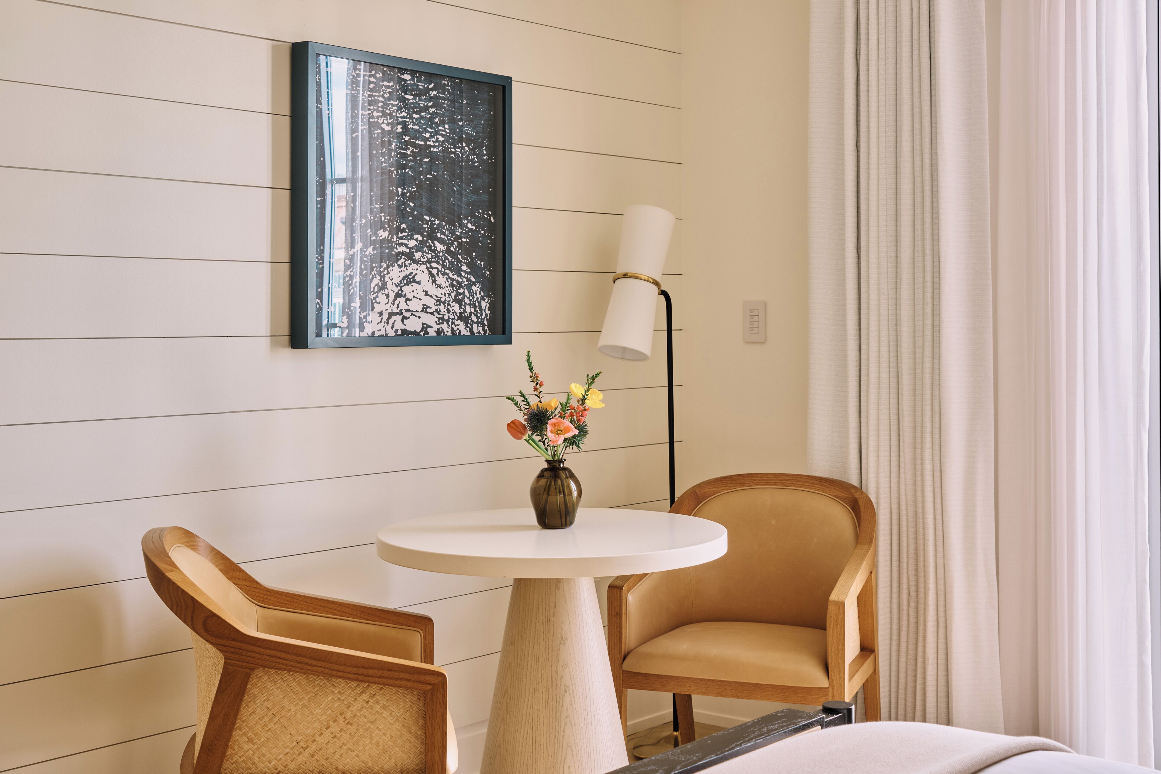 Hotelkamer met een ronde tafel, twee stoelen, een vaas met bloemen, een lijst met kunstwerk, een vloerlamp en een raam met gordijnen.