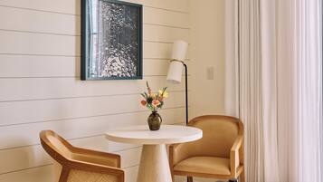Hotelkamer met een ronde tafel, twee stoelen, een vaas met bloemen, een lijst met kunstwerk, een vloerlamp en een raam met gordijnen.