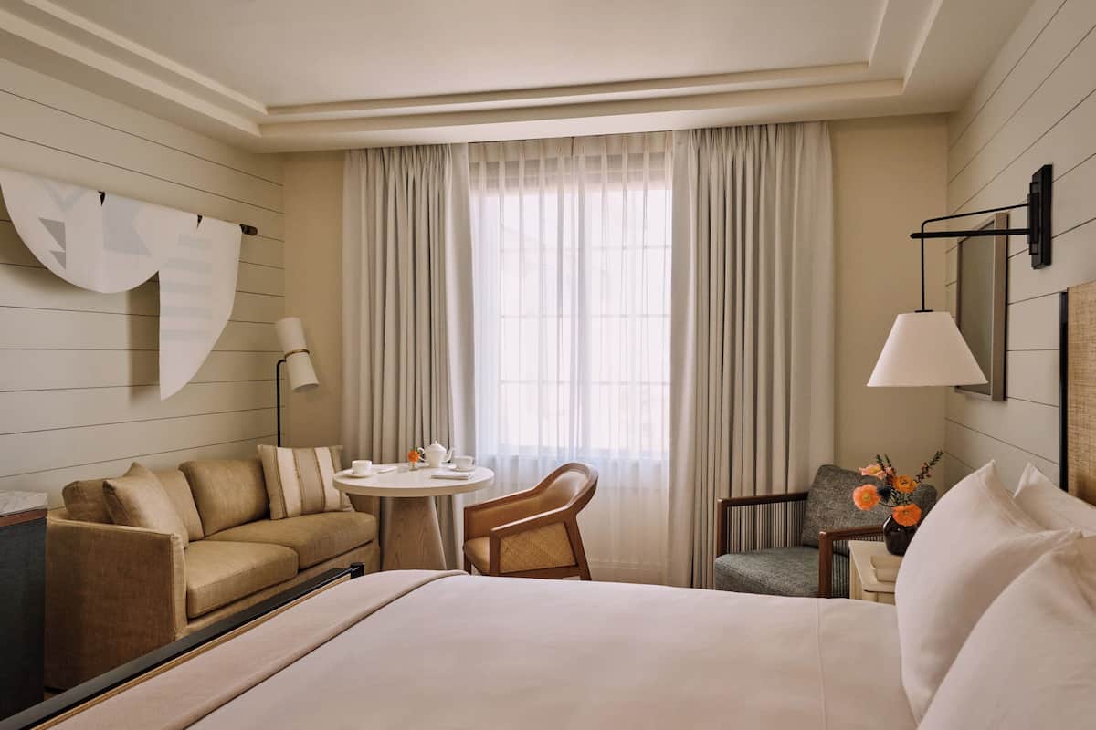A hotel room with a bed, a sofa, a small round table, two chairs, a lamp, and a window with curtains.