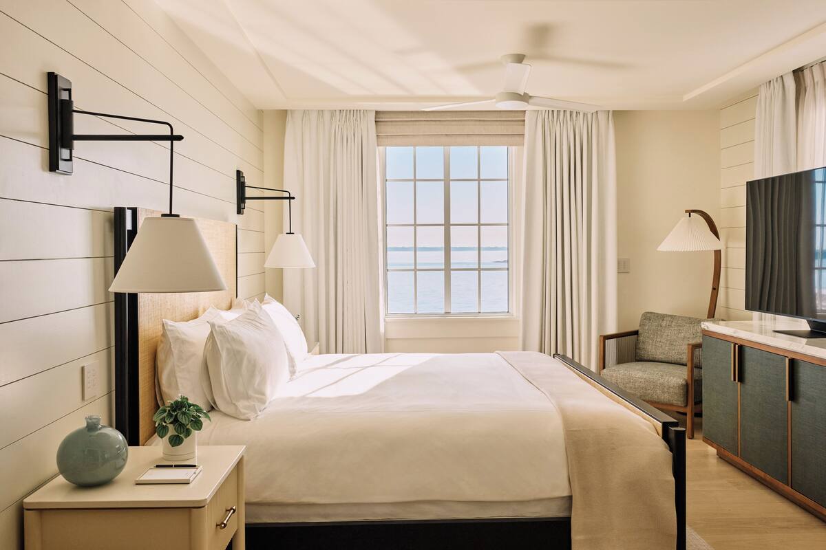 A hotel room with a bed, bedside tables, a desk, a chair, a TV, and a window with curtains.