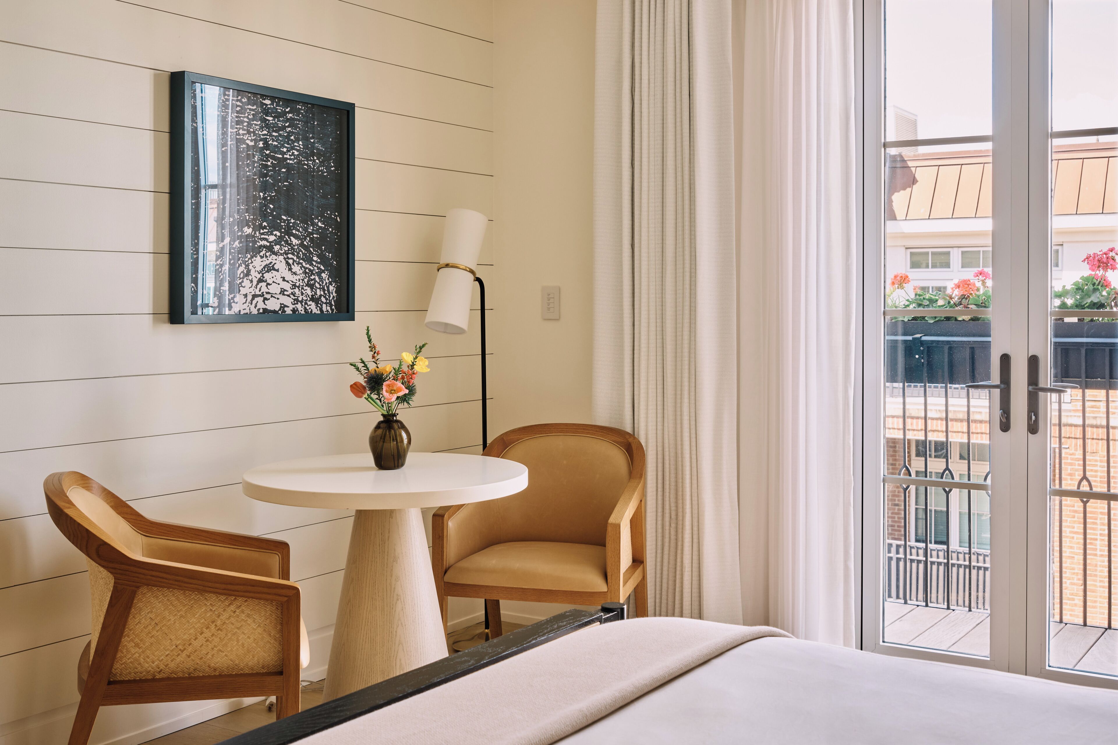 Hotelkamer met een bed, twee stoelen, een ronde tafel met een bloevas, een schilderij aan de muur en uitzicht op het balkon.