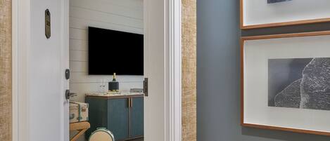 A hallway with a flat-screen TV mounted on the wall, a small table with a bottle and glasses, and a suitcase on the floor.