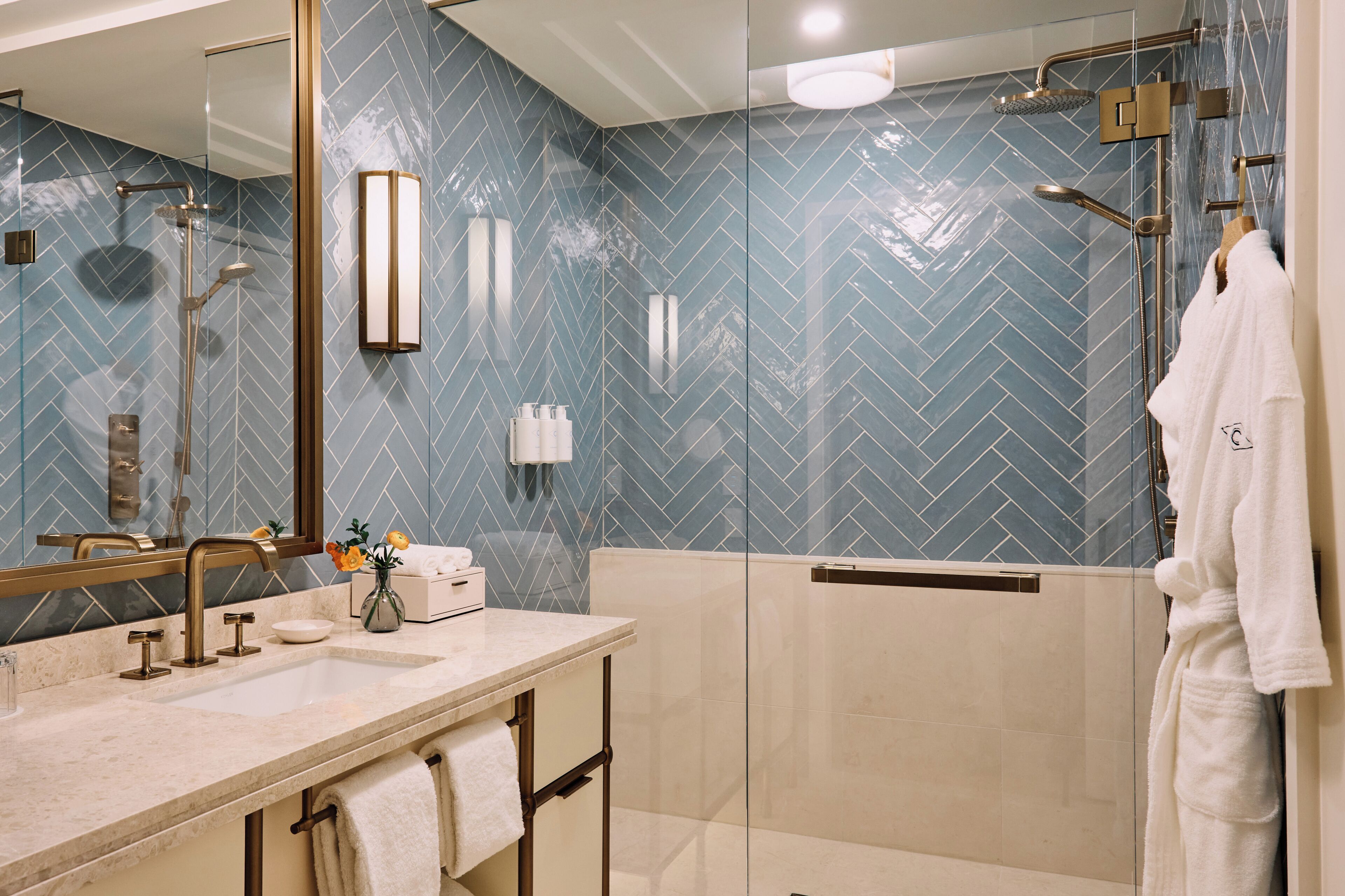 A modern bathroom with a herringbone tile backsplash, a large mirror, dual sinks, a glass shower enclosure, and a white robe hanging on the wall.