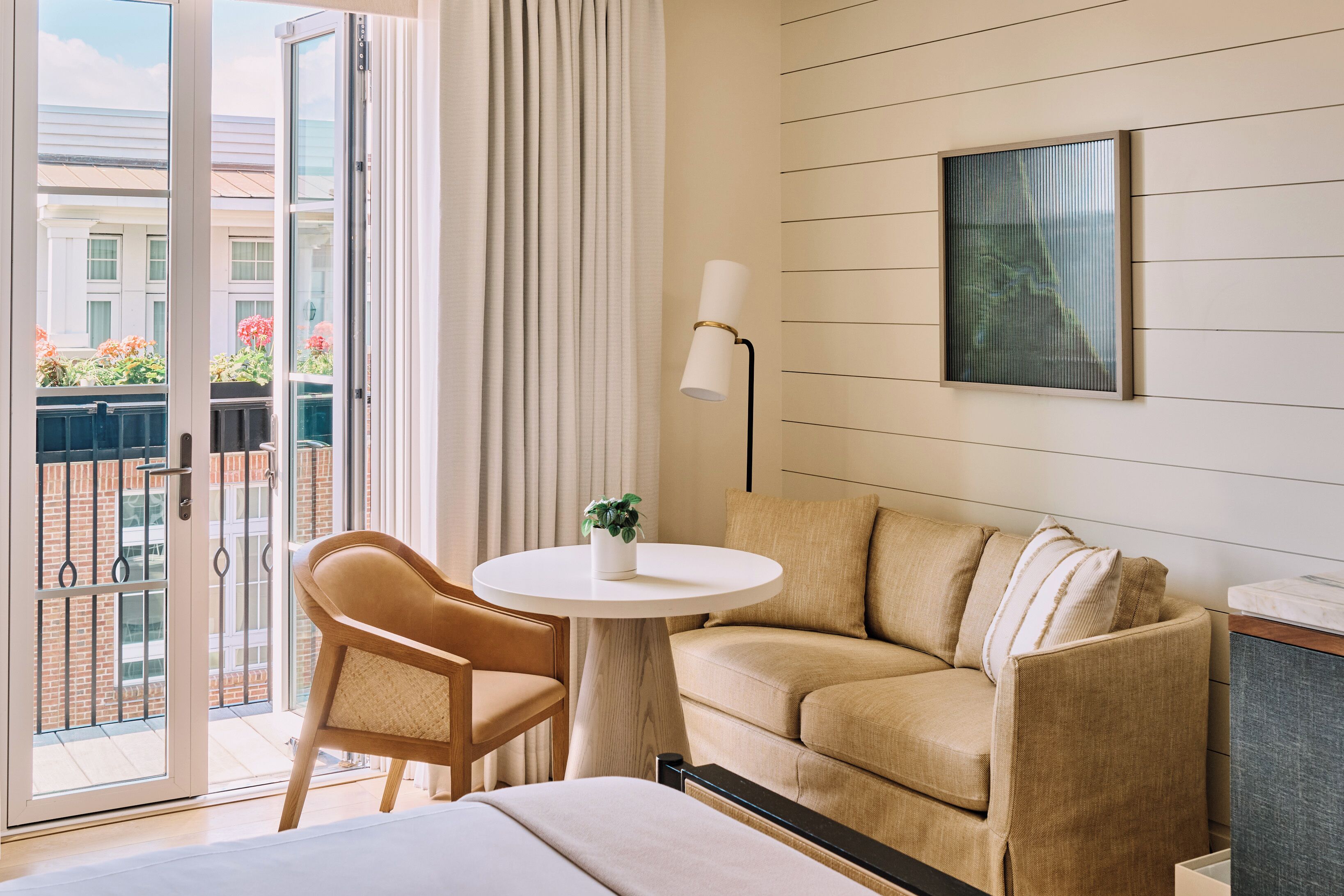 A hotel room with a sofa, a chair, a small table with a plant, a lamp, and a painting on the wall.