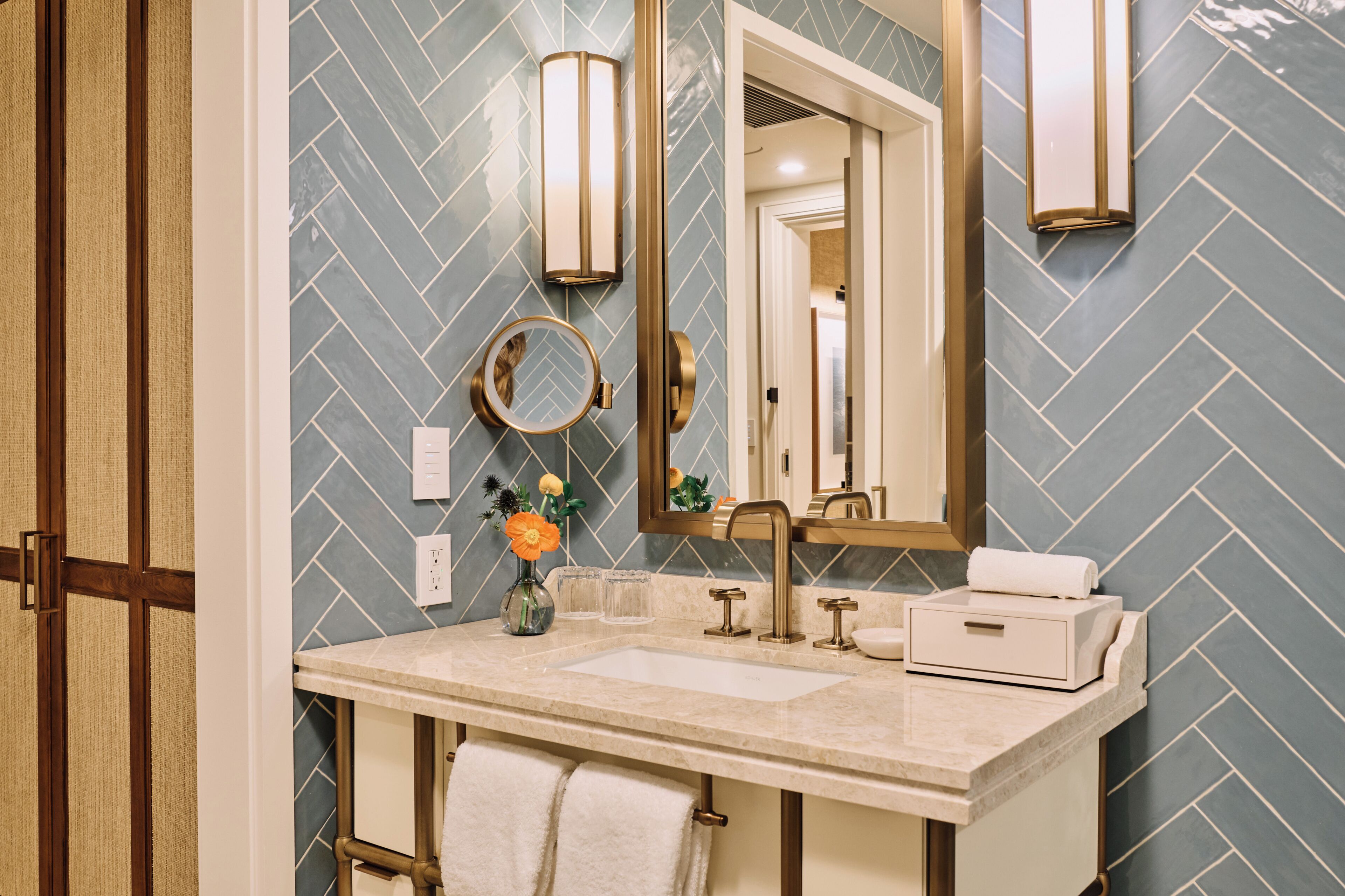 Un baño con pared de diseño en patrón de espiga, un espejo redondo, un florero con flores, una toalla y un lavabo con grifo.