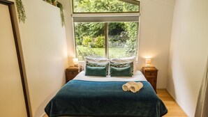 Een slaapkamer met een groot raam, een bed met een blauwe deken en groene kussens, twee nachtkastjes met lampen, en een paar opgerolde handdoeken op het bed.