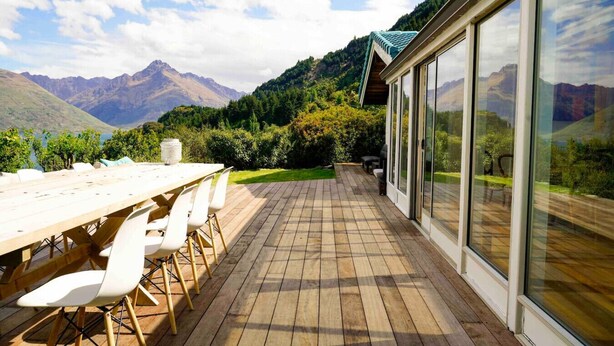 Un patio exterior con mesa y sillas de madera, grandes puertas correderas de vidrio y vistas a un paisaje montañoso.