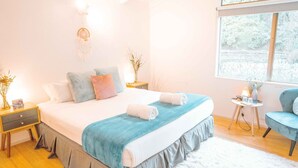 Un dormitorio con una cama grande, mesitas de noche, una pequeña mesita auxiliar, una silla y una ventana con vista a árboles.