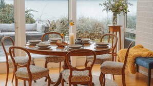 Un comedor con una mesa redonda dispuesta para seis personas, rodeada de sillas de madera con cojines estampados. Un florero con flores y una vela están sobre la mesa.