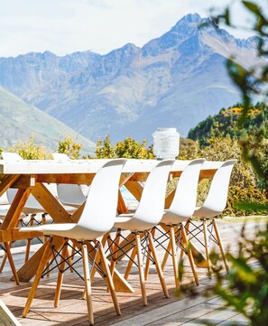 Une table à manger avec des chaises blanches et un pichet en verre, installée à l’extérieur, avec une vue sur les montagnes en arrière-plan.