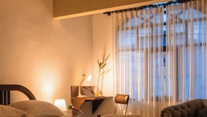 Quarto com cama, escrivaninha com laptop, cadeira e janela com cortinas leves.