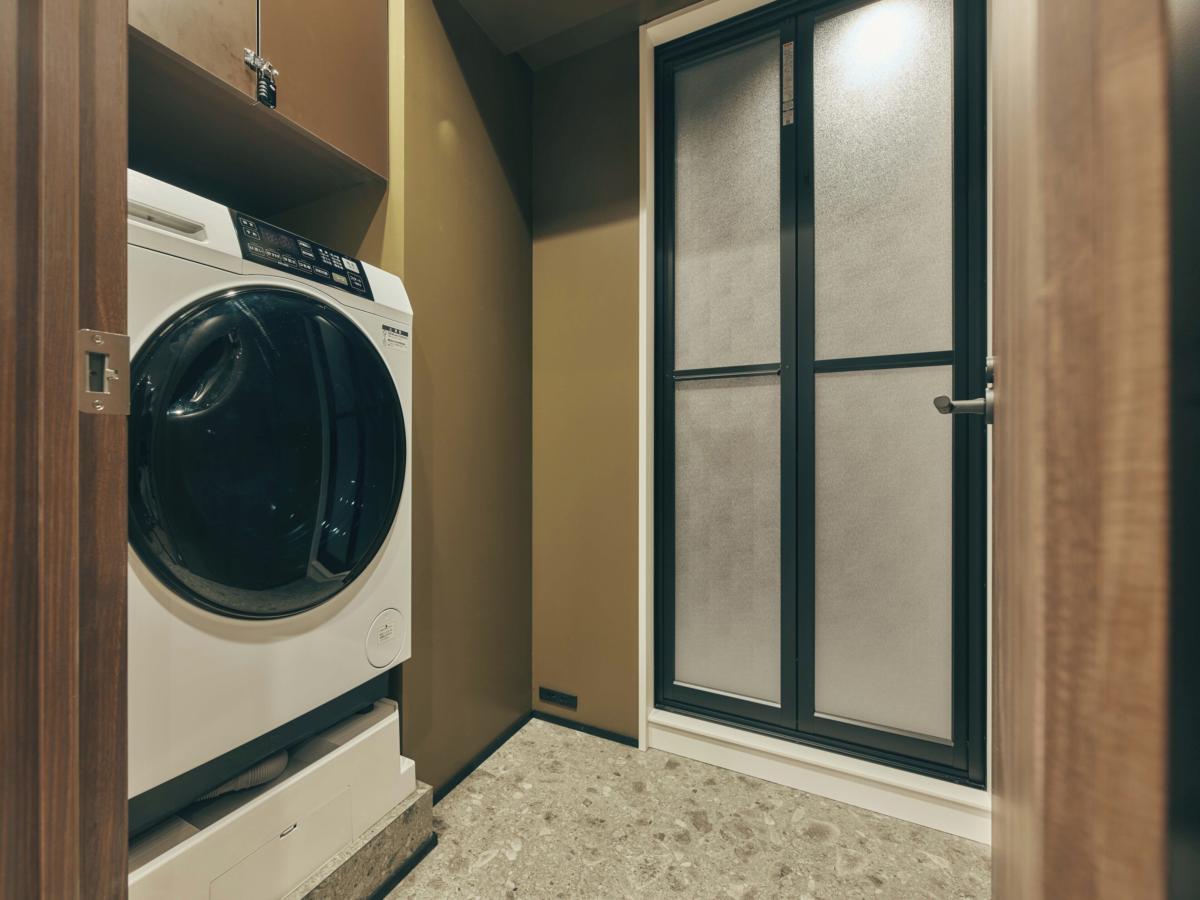 A laundry room with a front-loading washing machine, a door with frosted glass panels, and a beige tiled floor.