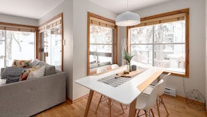 Un espace repas moderne avec une table blanche, des chaises en bois et un luminaire suspendu. De grandes fenêtres à cadre en bois laissent entrer la lumière naturelle et offrent une vue sur l’extérieur.
