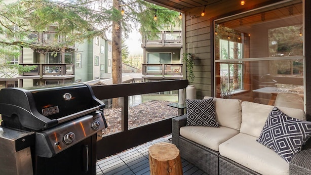 Un espace extérieur abrité, équipé d'un barbecue, d'un canapé en osier et de grandes fenêtres donnant sur un quartier résidentiel.