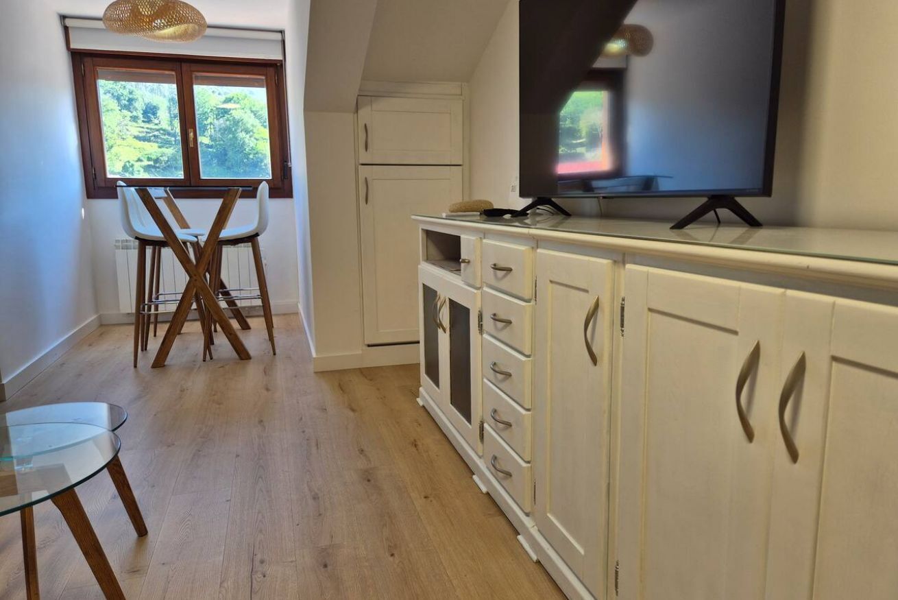 Una cocina con televisión, gabinetes blancos, una mesa de comedor con sillas y una ventana con vistas a zonas verdes.