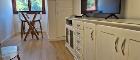 Una cocina con televisión, gabinetes blancos, una mesa de comedor con sillas y una ventana con vistas a zonas verdes.