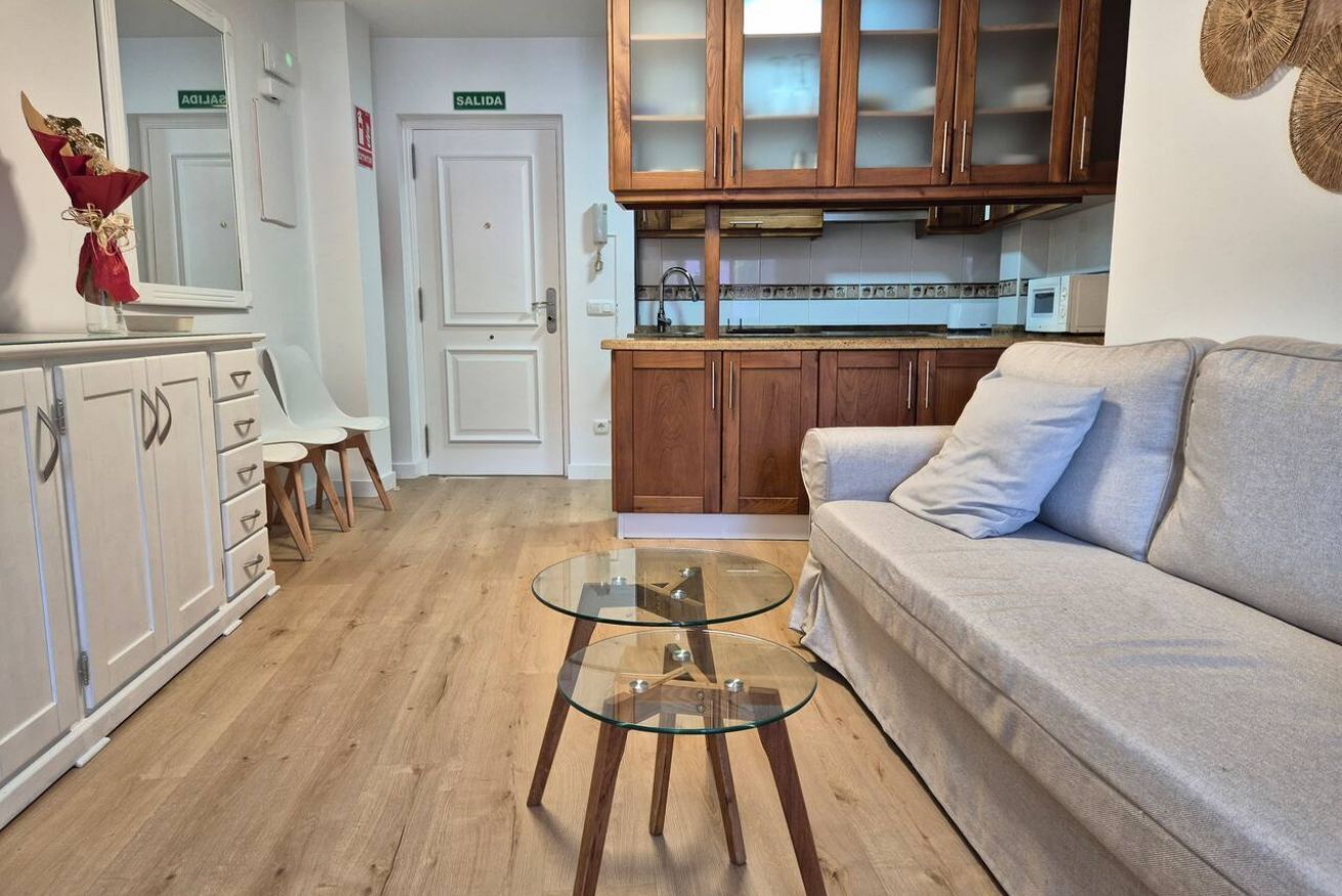 Een woonkamer met een glazen salontafel, een bank, houten kasten en een kitchenette.