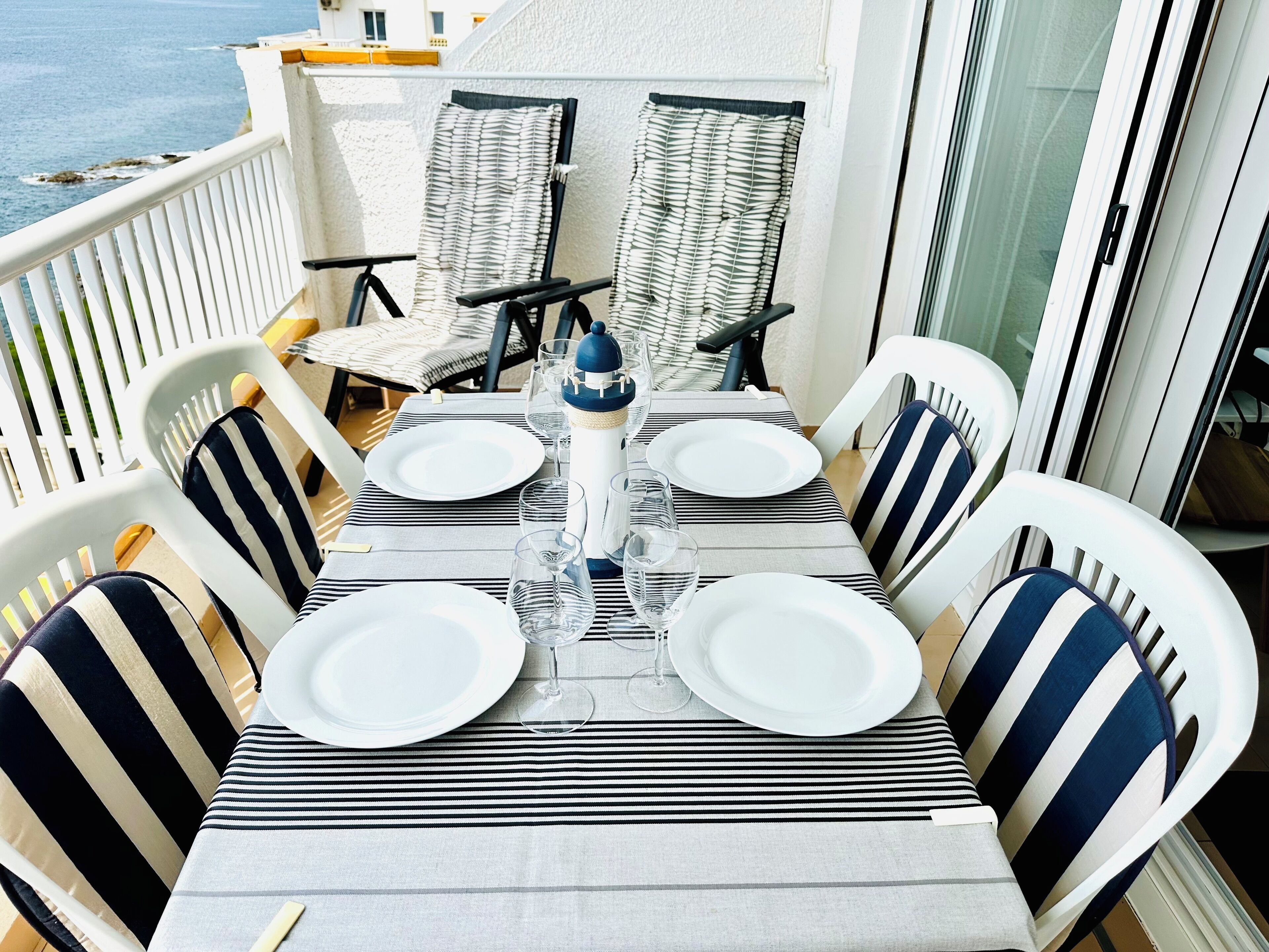 Uma área de refeições com uma mesa posta para quatro pessoas, rodeada por cadeiras listradas, um farol como peça central e vista para o mar.