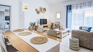 Ein modernes Wohnzimmer mit Esstisch aus Holz, bequemen Sitzgelegenheiten, einem Fernseher und Blick ins Freie.