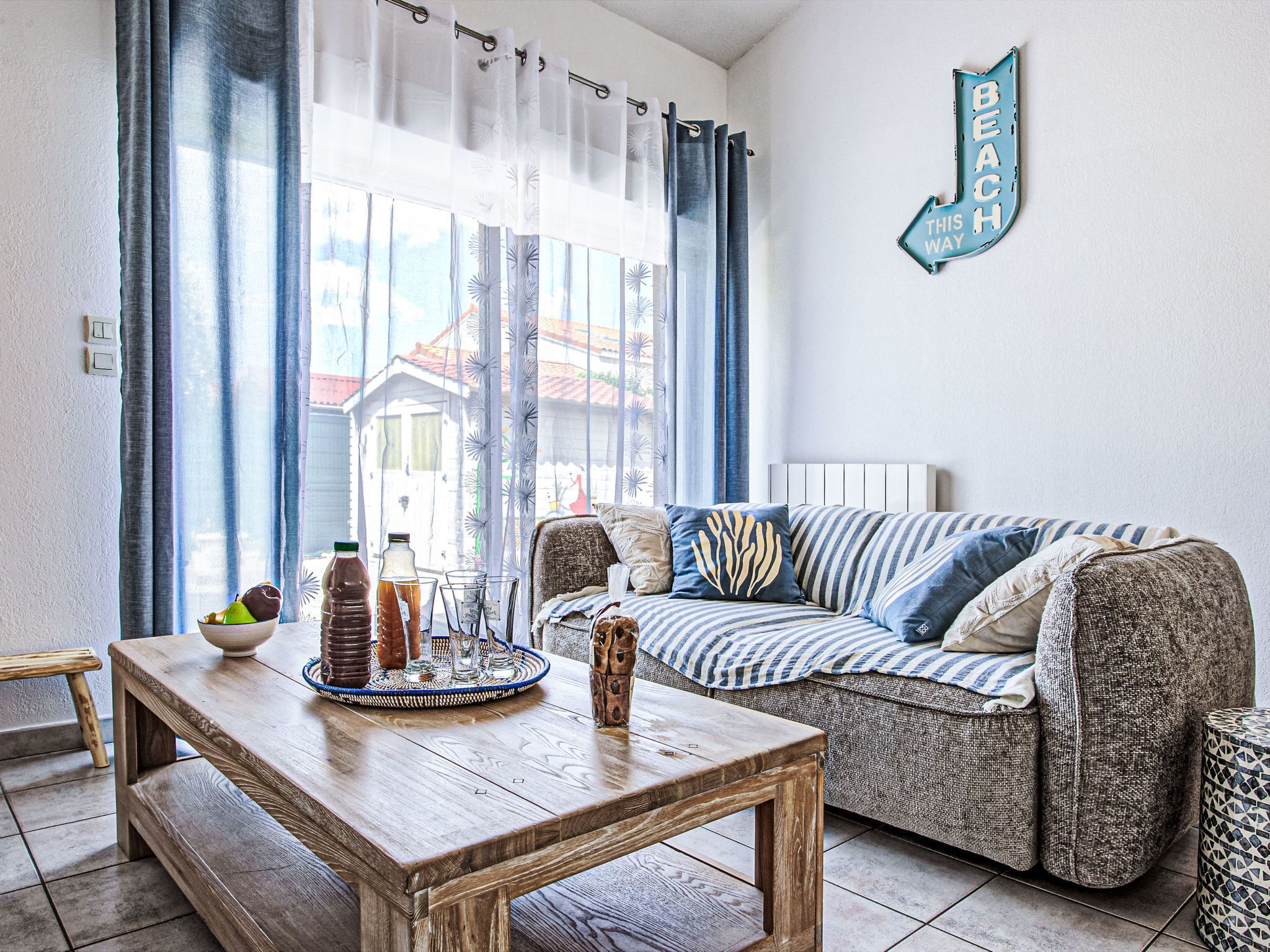 Un salon avec une table basse en bois, un canapé à rayures et une vue sur une maison à travers des rideaux transparents.
