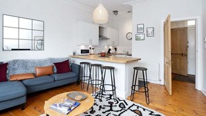 Un salon moderne avec un canapé, une table basse et des tabourets de bar. On aperçoit une cuisine dotée d’un îlot central et de tabourets de bar.