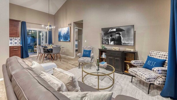 A modern living room with a grey sofa, patterned armchairs, a coffee table, and a flat-screen TV. There is a dining area with a table and chairs, and a kitchen visible through an open door. - Blue Oasis with Private Heated Pool at NO COST (Tempe)