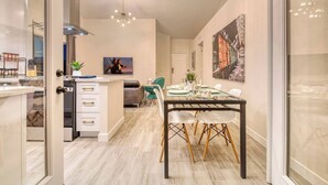 A modern kitchen with a glass door, a dining area with a glass table, and a living space with a sofa and a TV.