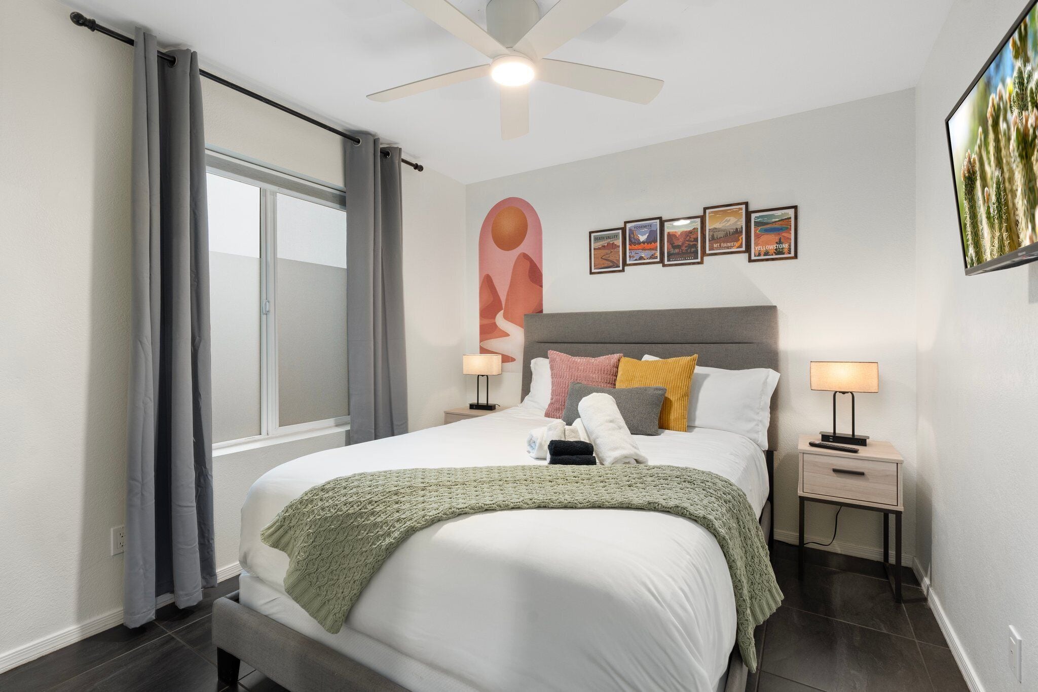 A modern bedroom with a bed, two bedside tables, a ceiling fan, a window with curtains, and wall art.