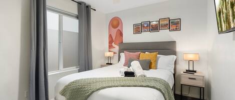 A modern bedroom with a bed, two bedside tables, a ceiling fan, a window with curtains, and wall art.