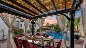 An outdoor patio with a pool, red chairs, a table set for dining, and a pergola with a thatched roof. - Fabulous entertainers Paradise! Pool & Spa (Phoenix)